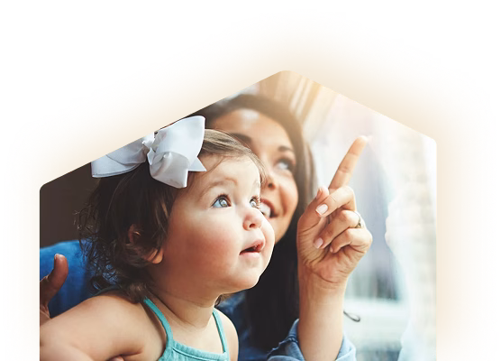 A mother points toward something outside a bright window as her young daughter, wearing a white bow, watches with wonder in a tender moment.