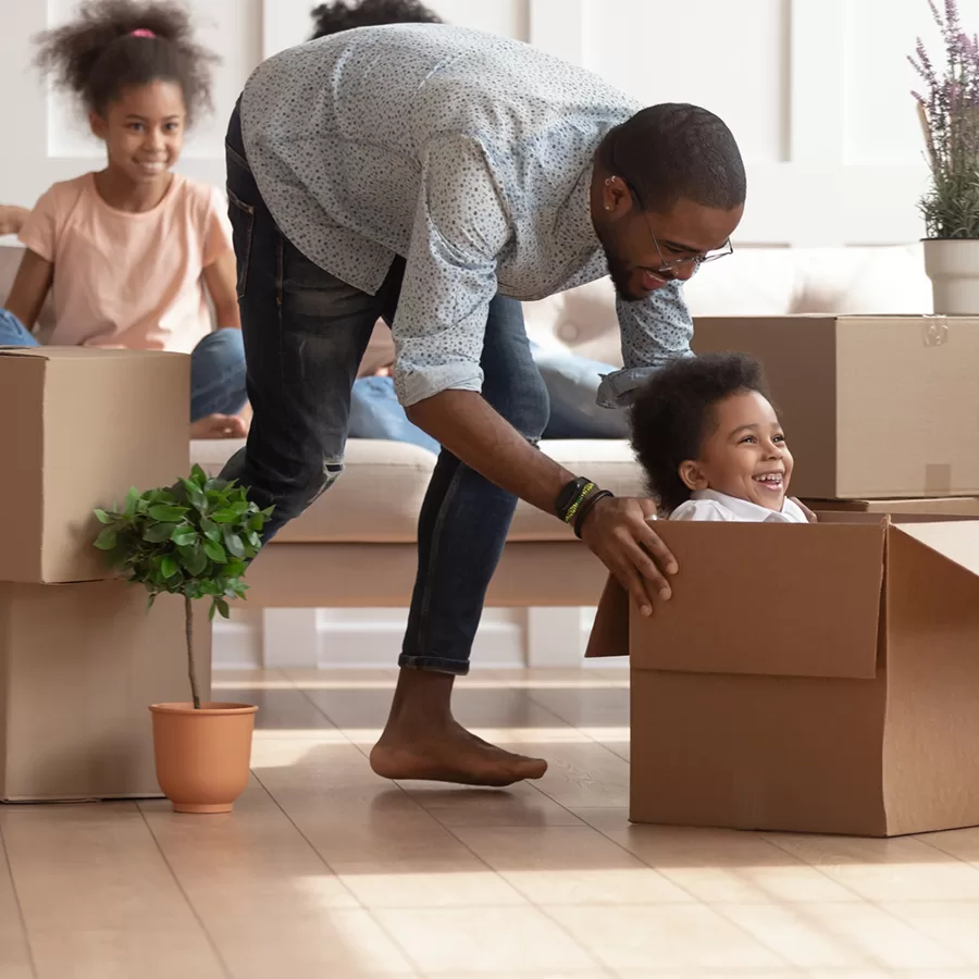 A smiling father playfully steadies a child sitting in a cardboard box during a family move, while another child watches from the sofa in a bright living room.