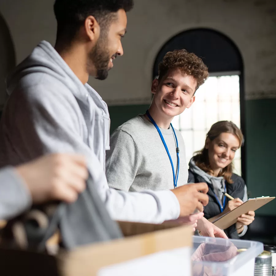 A diverse group of students smiles and talks around a classroom table, sorting materials into a clear bin; the collaborative scene feels warm and upbeat.