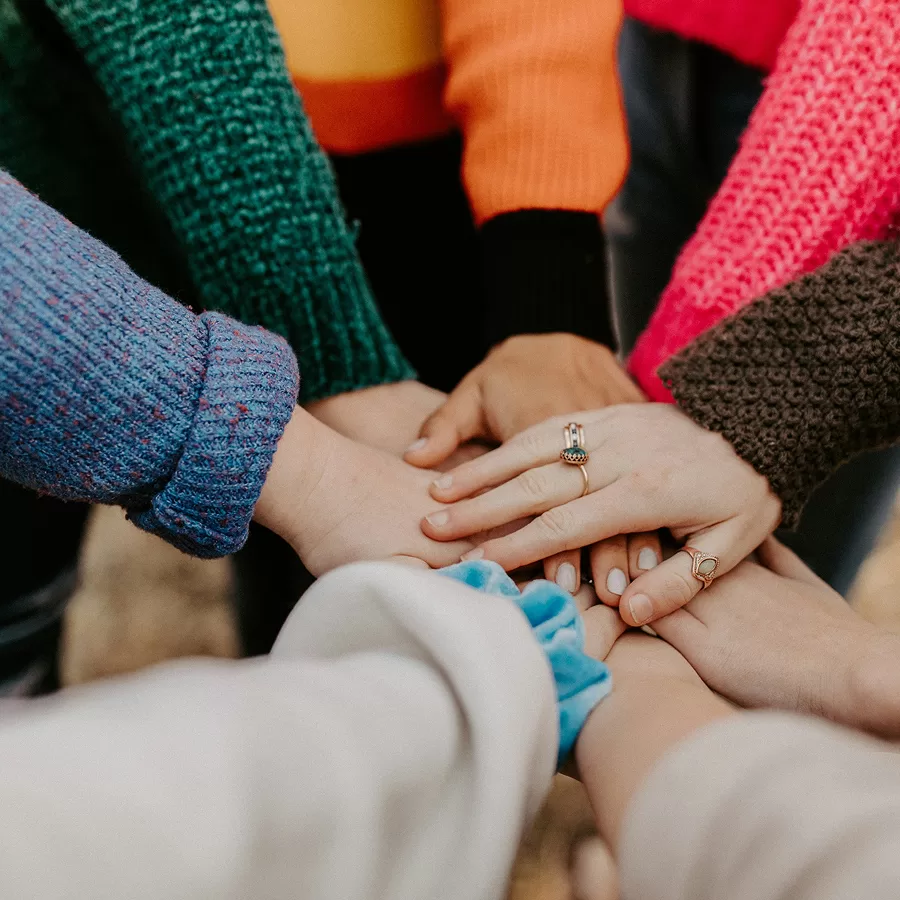 A diverse group stacks hands together, colorful sweater sleeves and rings visible, conveying teamwork, unity, and warm camaraderie.